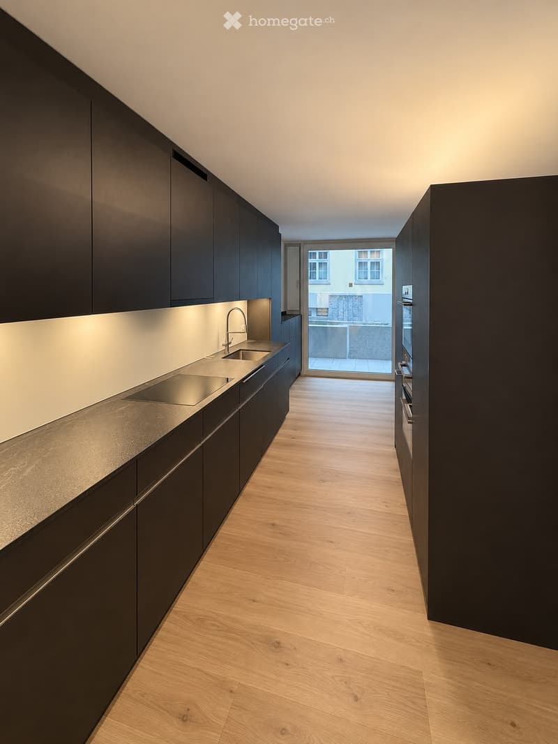 Modern kitchen with dark cabinets, light countertops, and a sleek sink area. The space features wooden flooring and a view of a balcony through a large window.