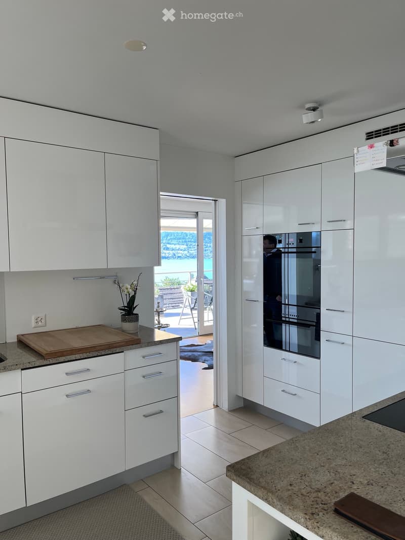 Modern kitchen with white cabinets, granite countertops, and a wooden cutting board on the counter. A vase with white flowers is placed on the counter, and an oven is visible in the background. The kitchen has a sliding glass door leading to a balcony wit