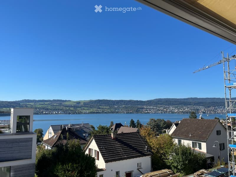 Balcony view overlooking a lake with surrounding hills, houses, and a clear blue sky.