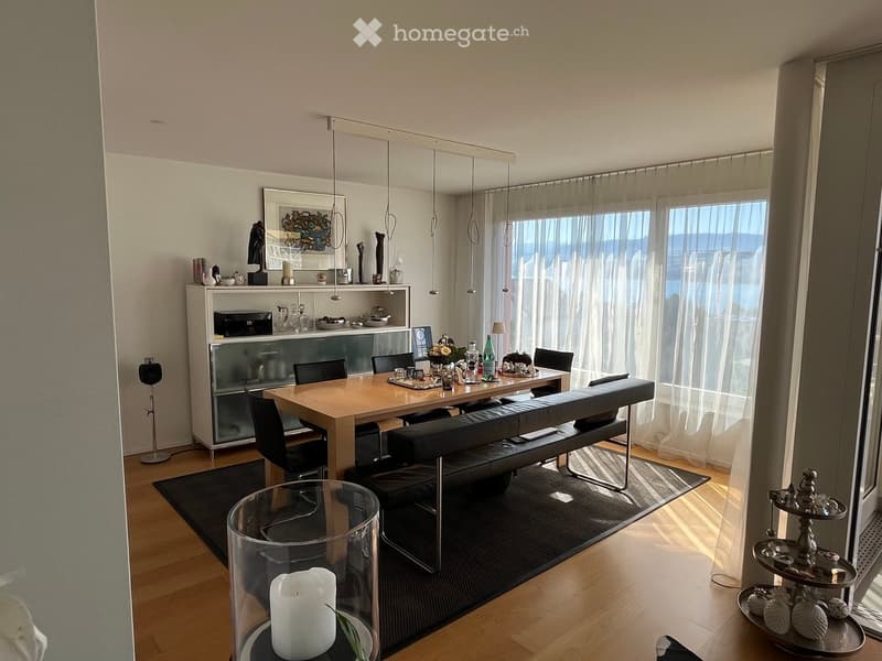 Modern dining area with a wooden dining table, black bench seating, chairs, and a glass display cabinet. Large windows with sheer curtains offer a view of the water.