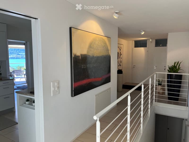 Modern hallway with white walls, tiled floor, staircase railing, and a large abstract painting on the wall. A view of a balcony and outdoor area is visible through an open doorway.