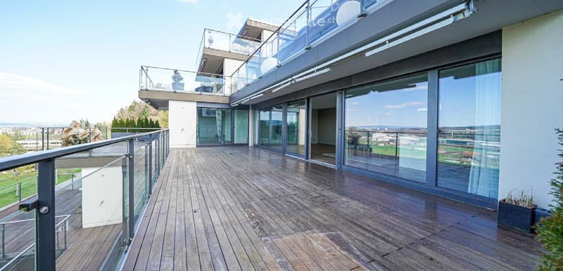 Spacious wooden deck with glass railings, offering a panoramic view of the surrounding landscape and cityscape.