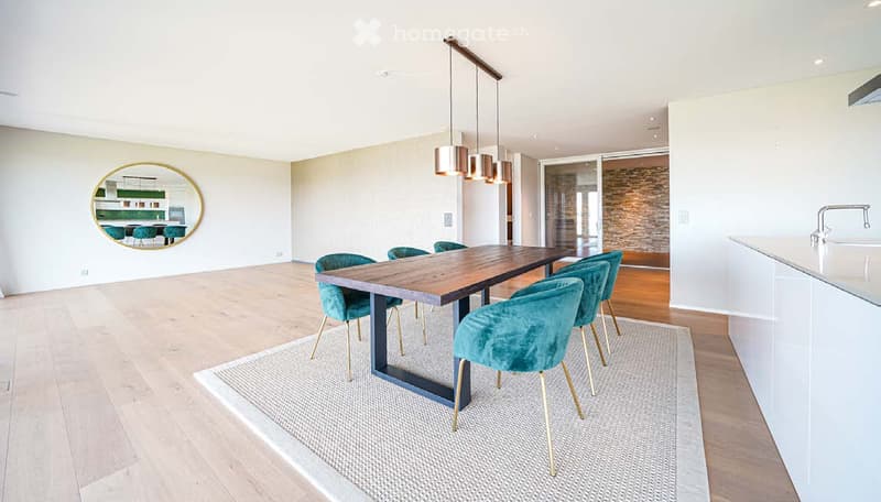 Dining area with a wooden table, teal upholstered chairs, a modern pendant light fixture, and a round mirror reflecting a kitchen area.