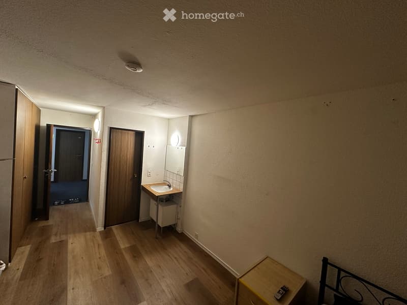 A narrow hallway with wooden flooring, beige walls, and recessed lighting. A small sink area is visible on the right, with a mirror, faucet, and cabinet. A door leads to another room on the left, and a staircase is visible at the end of the hallway.
