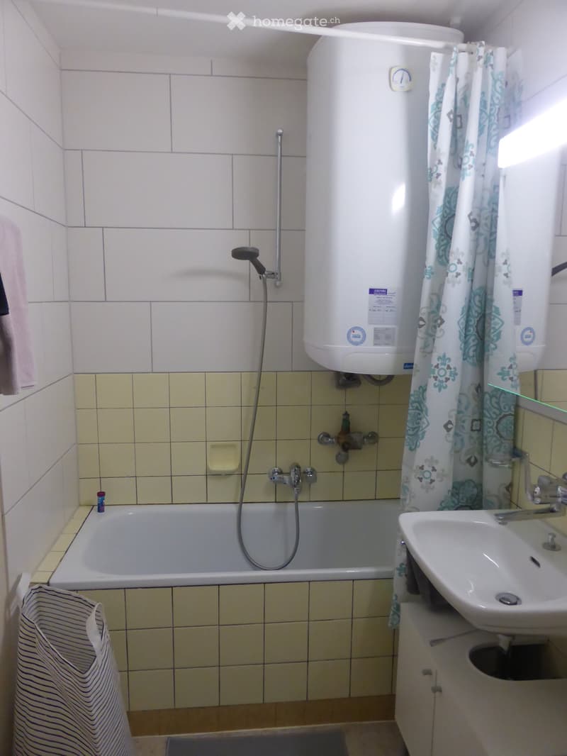 Bathroom with a bathtub, sink, showerhead, water heater, and a mirror. The walls are tiled, and there is a towel hanging on the side.