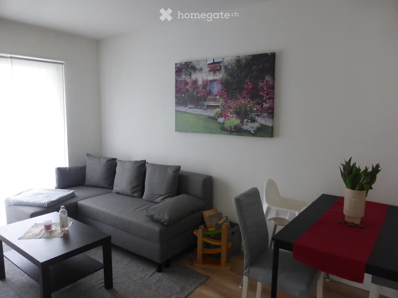 A modern living room with a grey sectional sofa, coffee table, dining table with chairs, and a potted plant on the table. The room features large windows with blinds and a large wall painting.