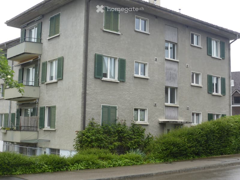 Four-story residential building with grey facade, green shutters, multiple windows, and balconies.