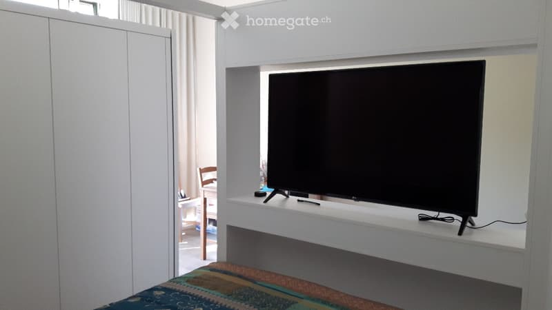 A modern, minimalistic room with a large flat-screen TV on a white shelf, a colorful patterned bedspread, and a small wooden chair and table visible in the background.
