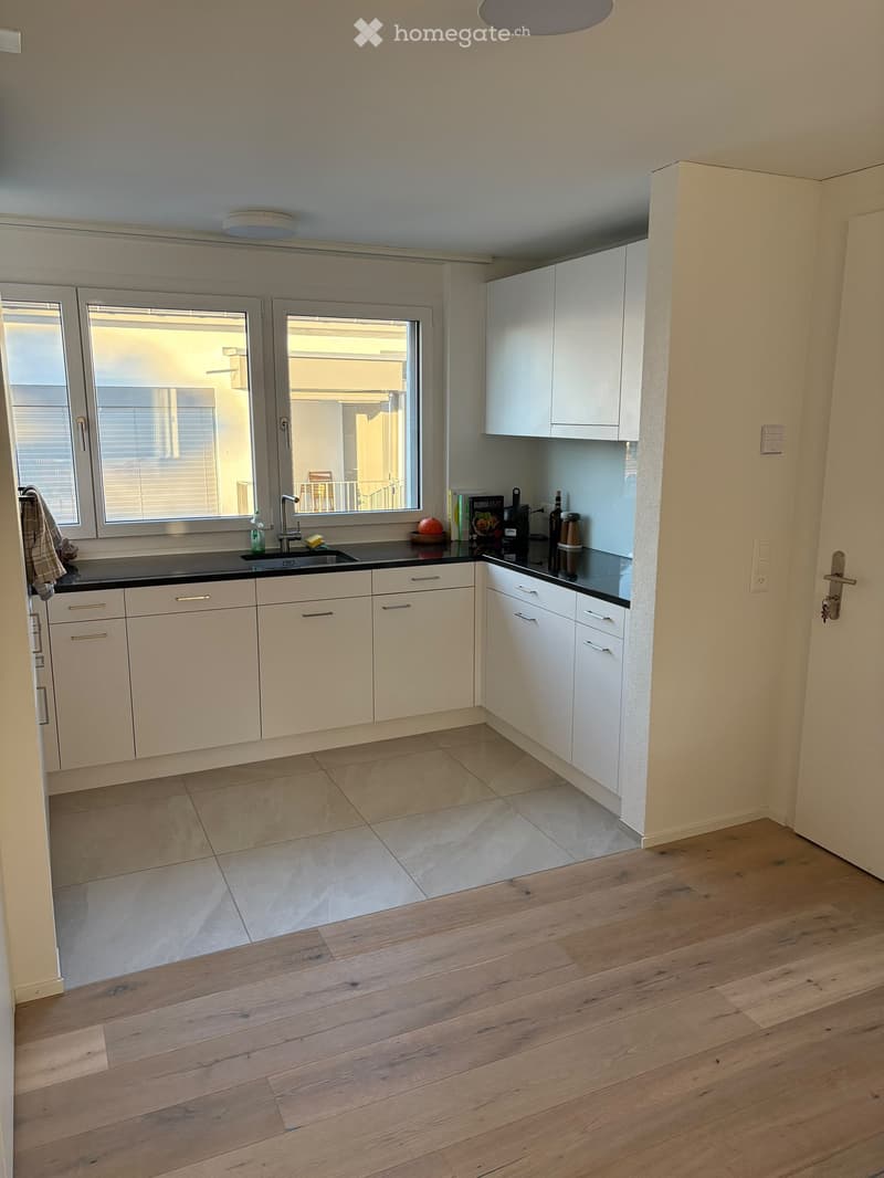 Modern kitchen with white cabinets, black countertops, and a large window overlooking a balcony. The floor is a mix of tiled and wooden surfaces.