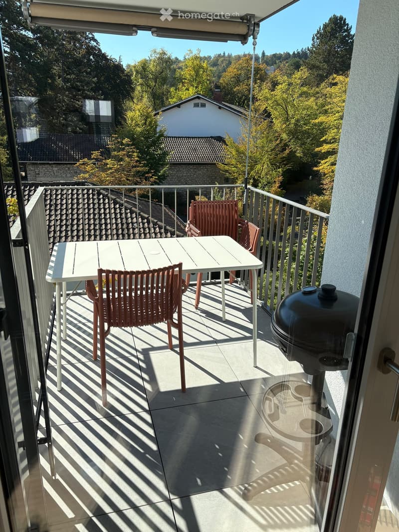 Balcony with a white table, four pink chairs, and a black grill, overlooking a tree-lined neighborhood.