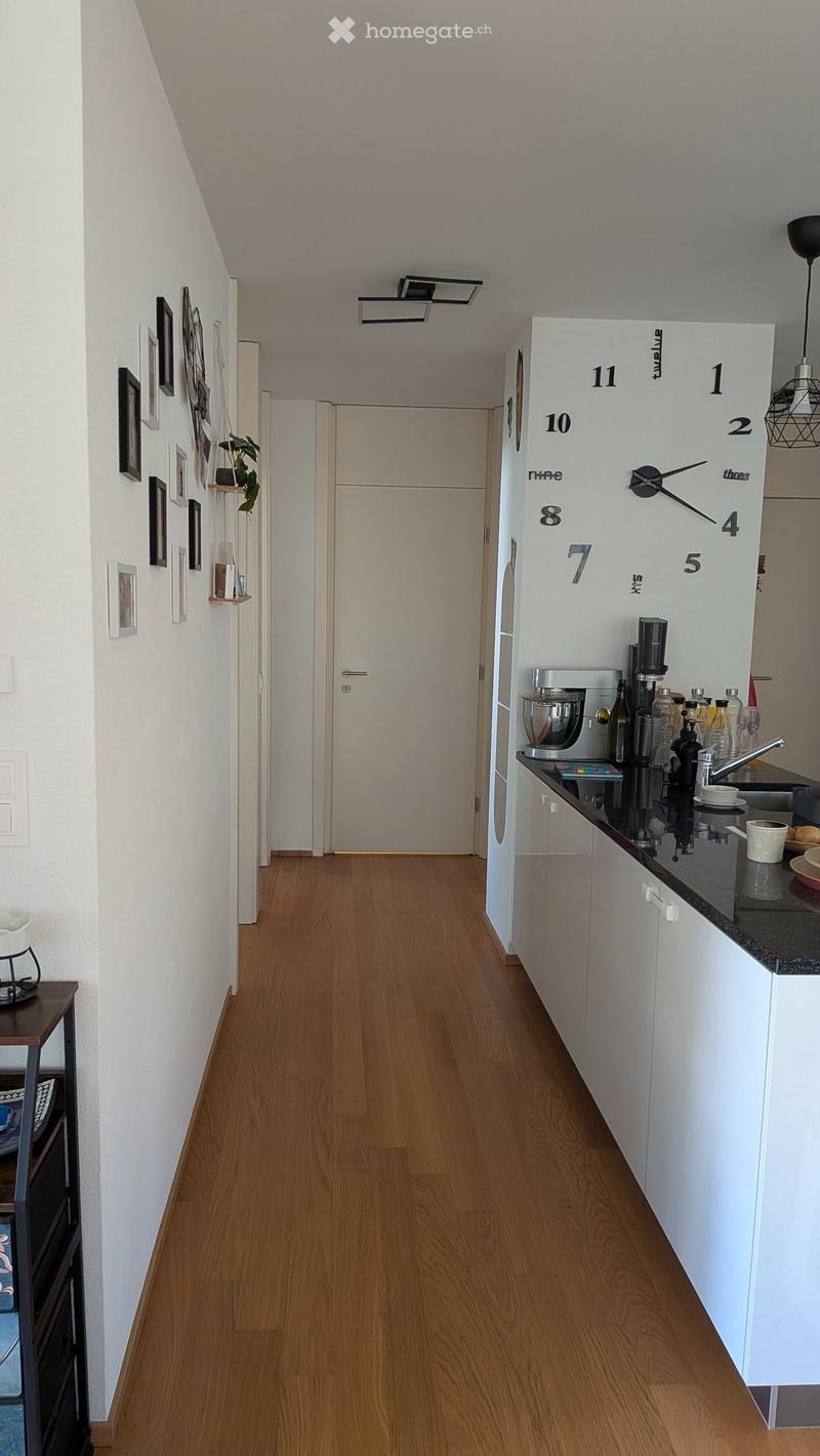 A narrow hallway with wooden flooring, white walls, and a small kitchen area visible on the right side. The kitchen features white cabinets, a countertop with various appliances, and a clock on the wall. A door is visible at the end of the hallway.