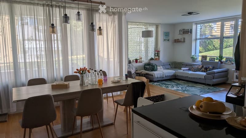 Bright living and dining area with a grey sectional sofa, wooden dining table with chairs, pendant lights, and large windows with sheer curtains.