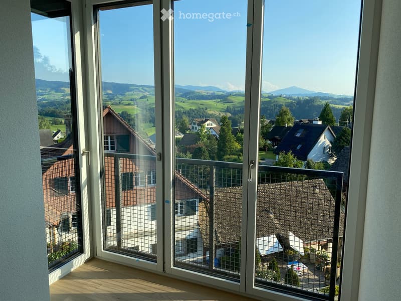 Balcony with metal railing, overlooking a scenic countryside with houses and distant mountains.