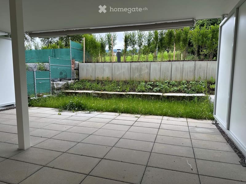 A modern patio area with large concrete tiles, a raised garden bed with plants and grass, and a concrete wall in the background.