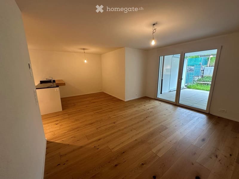 Empty room with wooden flooring, white walls, and a sliding glass door leading to a patio. A small kitchen area with a sink and countertop is visible on the left side.