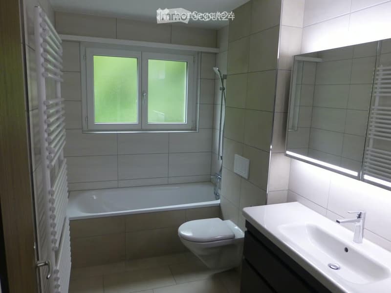 Modern bathroom with a bathtub, toilet, sink, mirror, and radiator. The room features tiled walls and a window with blinds.