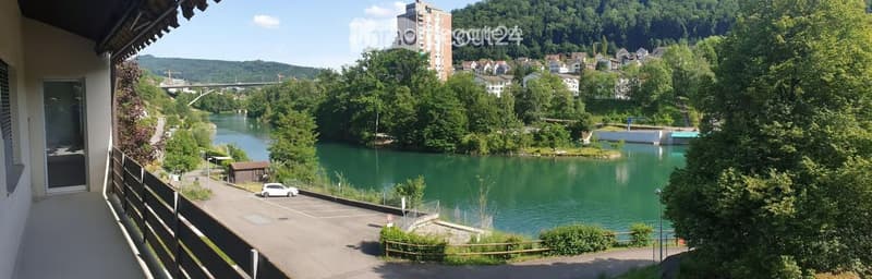 Balcony overlooking a river with a town and hills in the background, parking area nearby.