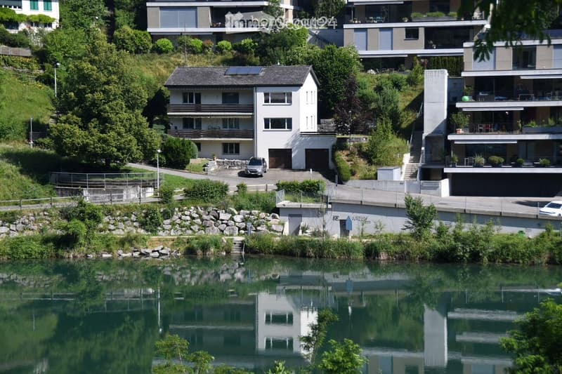 A modern residential complex situated on a hillside overlooking a calm river, featuring multiple buildings with balconies, large windows, and a parking area.