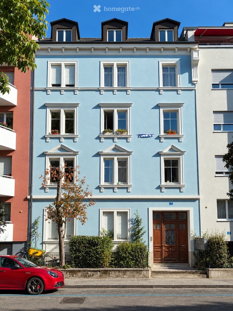 A three-story residential building painted in light blue with white trim around the windows and roofline. The building features multiple windows with shutters, small balconies with potted plants, and a wooden front door. A red car is parked on the street