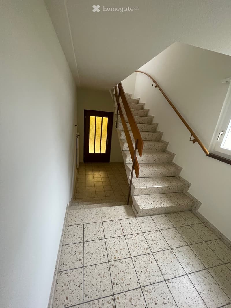 A narrow hallway with tiled floors and walls, featuring a staircase with a wooden handrail leading upwards, and a door with frosted glass panels at the end of the corridor.