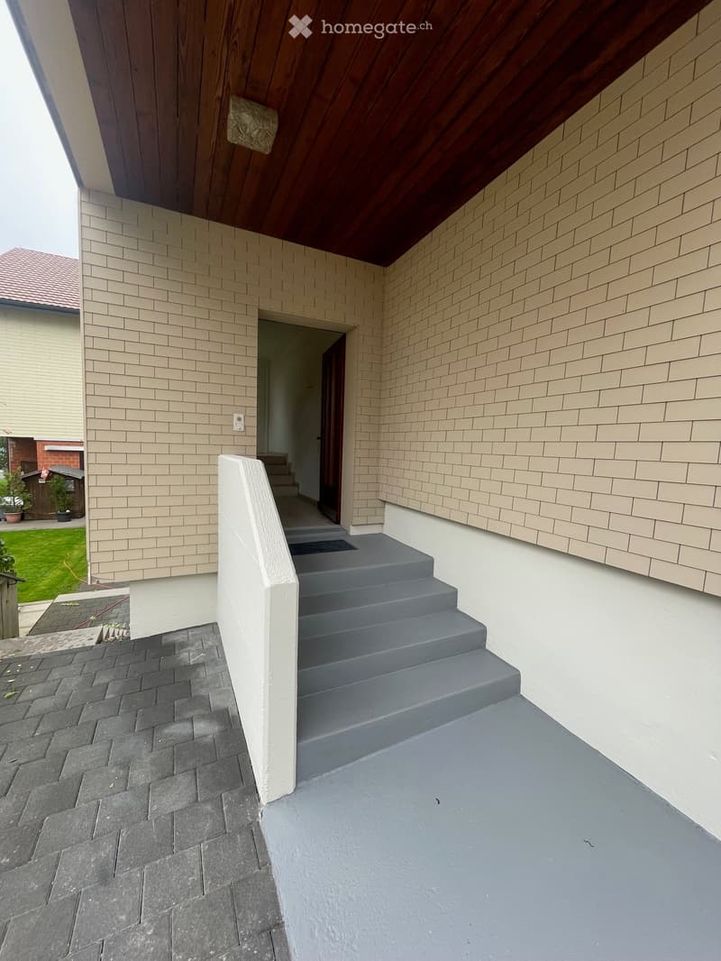 Modern balcony with grey concrete steps, grey tiled floor, and beige brick walls. Wooden roof overhang visible.