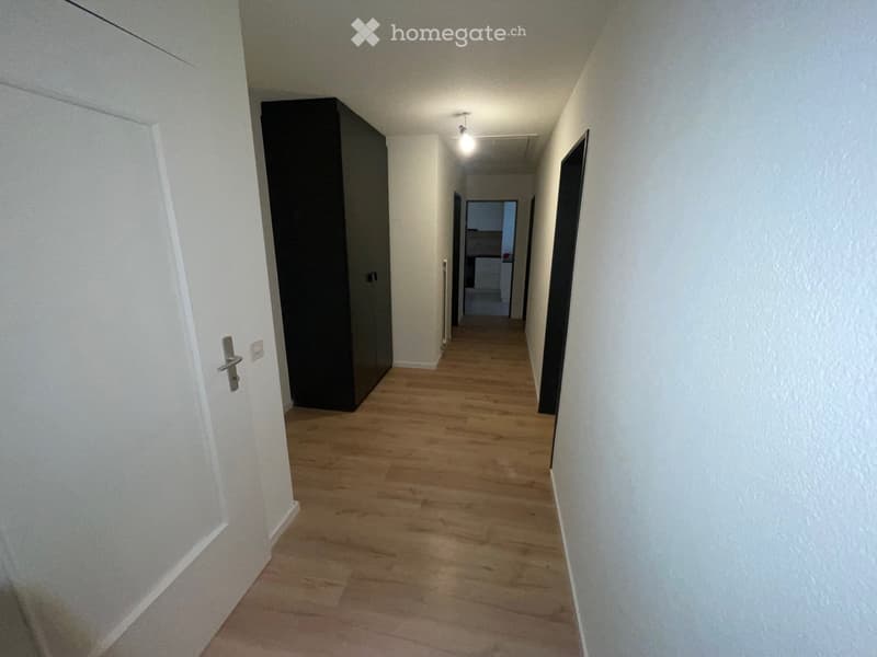 Empty hallway with wooden flooring, white walls, and a closed door on the left. A dark-colored cabinet is visible against the wall.