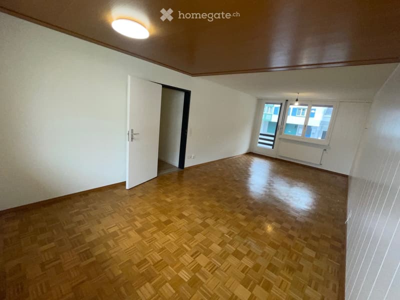 Empty room with wooden flooring, white walls, and a large window with a view of neighboring buildings.