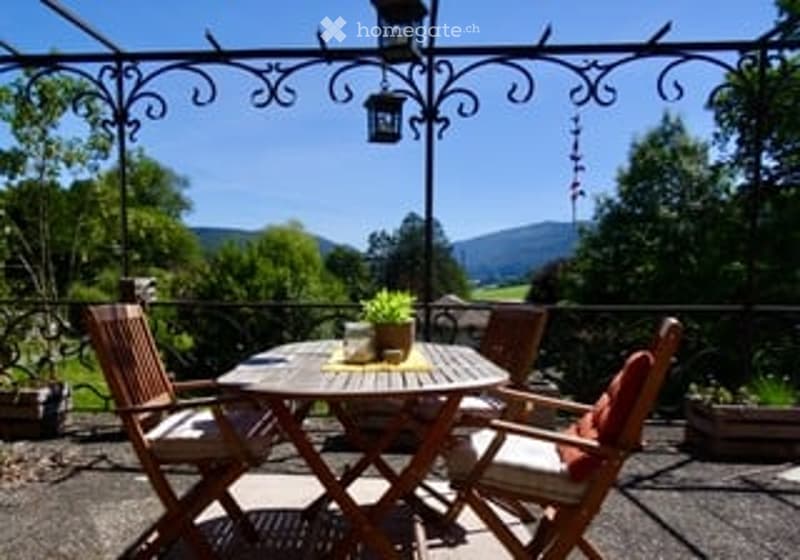 Wooden patio furniture set including a round table and four chairs, with a potted plant on the table, surrounded by a metal pergola with hanging lanterns.