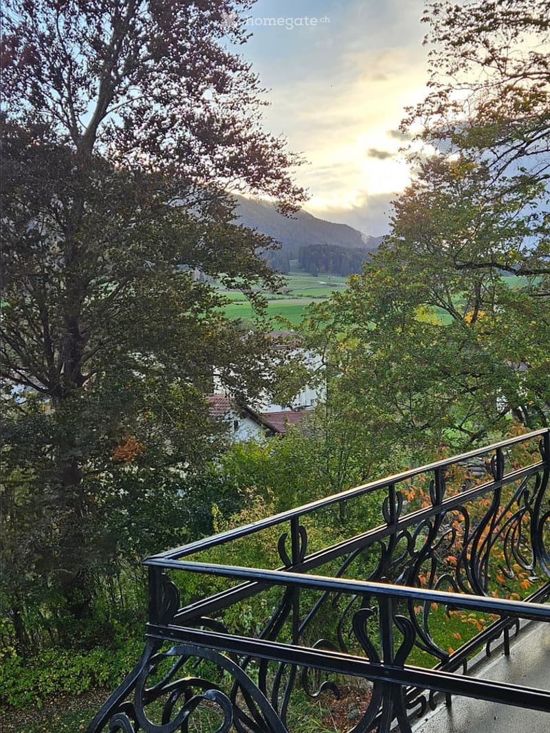 Balcony with ornate metal railing overlooking a scenic valley, trees, and distant mountains.