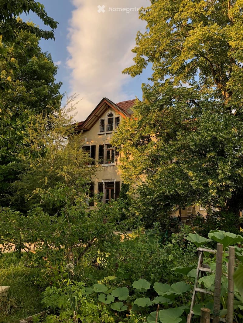 A lush garden with dense foliage and a small house partially visible behind the greenery. The house has a sloped roof and multiple windows, surrounded by tall trees and plants.