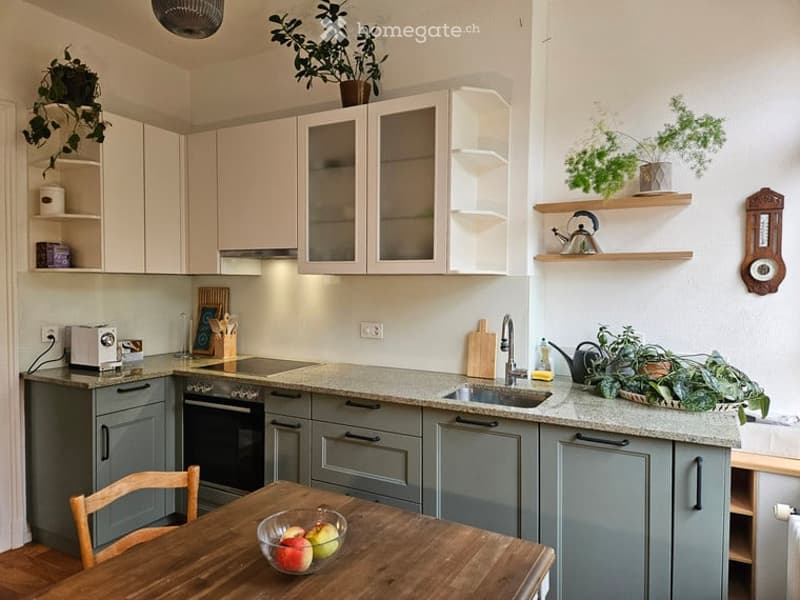 Modern kitchen with light-colored cabinets, grey lower units, a wooden dining table with a bowl of fruit, and various appliances including a toaster, oven, and sink.