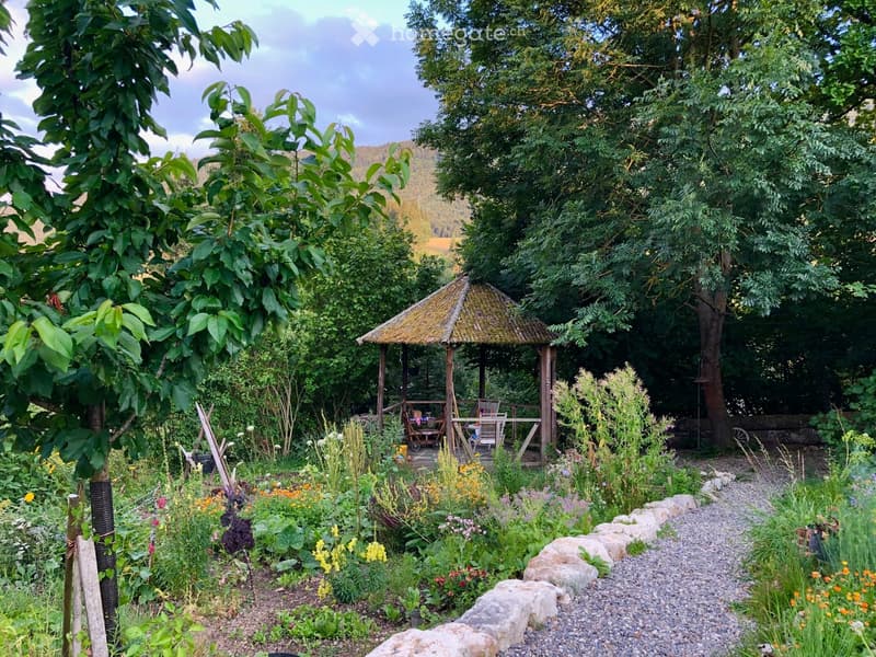 A lush garden with a thatched-roof gazebo, colorful flowers, trees, and a stone pathway.