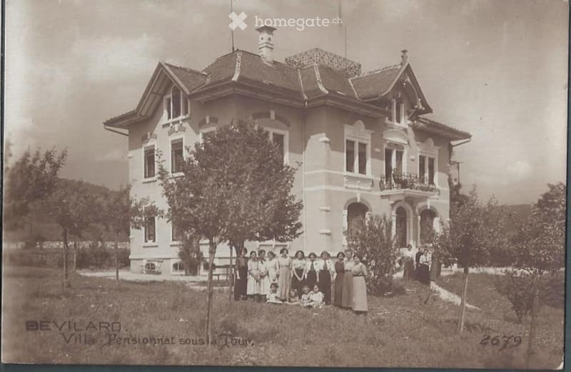 A large, two-story villa with a pitched roof, multiple windows, and decorative architectural details. The building is surrounded by a grassy area with trees, and a group of people is gathered in front of it.