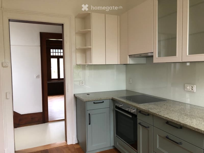 Modern kitchen with light grey cabinets, granite countertop, built-in oven, and induction cooktop. Open shelving and a glass backsplash are visible.