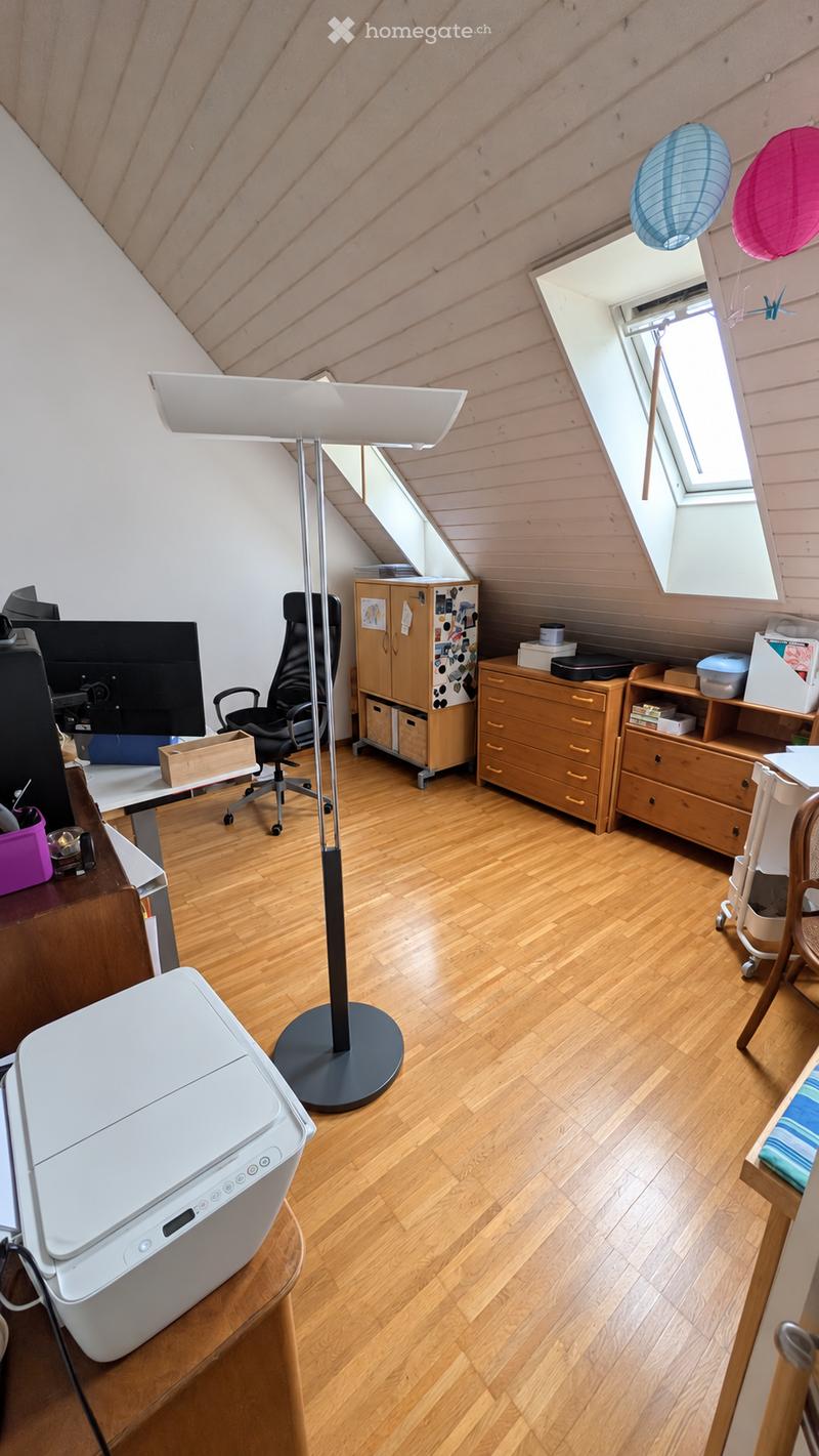 A well-lit attic room with wooden flooring, featuring a desk with a chair, a large screen, a printer, a tall floor lamp, and wooden storage units including cabinets and shelves.