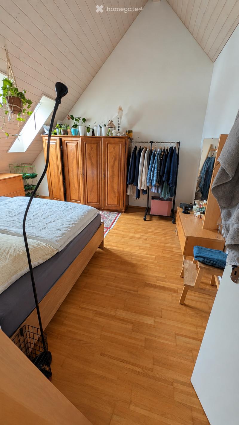 Cozy attic bedroom with wooden floors, slanted wooden ceiling, large window, bed, wardrobe, mirror, small table, and hanging plants.
