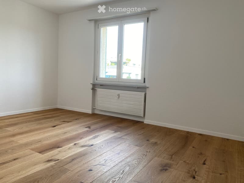 Empty room with wooden flooring, white walls, and a large window with a radiator below it.