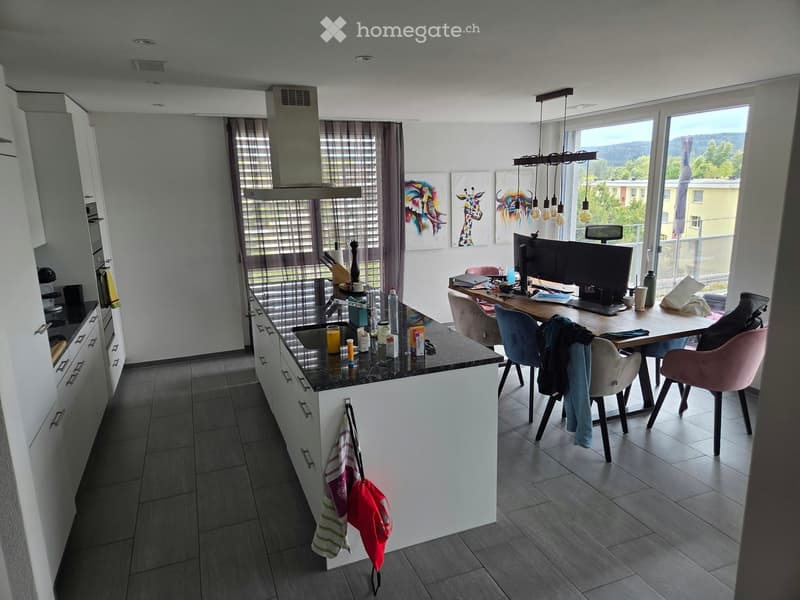 Open-plan kitchen and dining area with modern white cabinets, a central island, and a dining table with chairs. The kitchen features a sink, oven, microwave, and refrigerator. A laptop is placed on the dining table, and various kitchen items are scattered