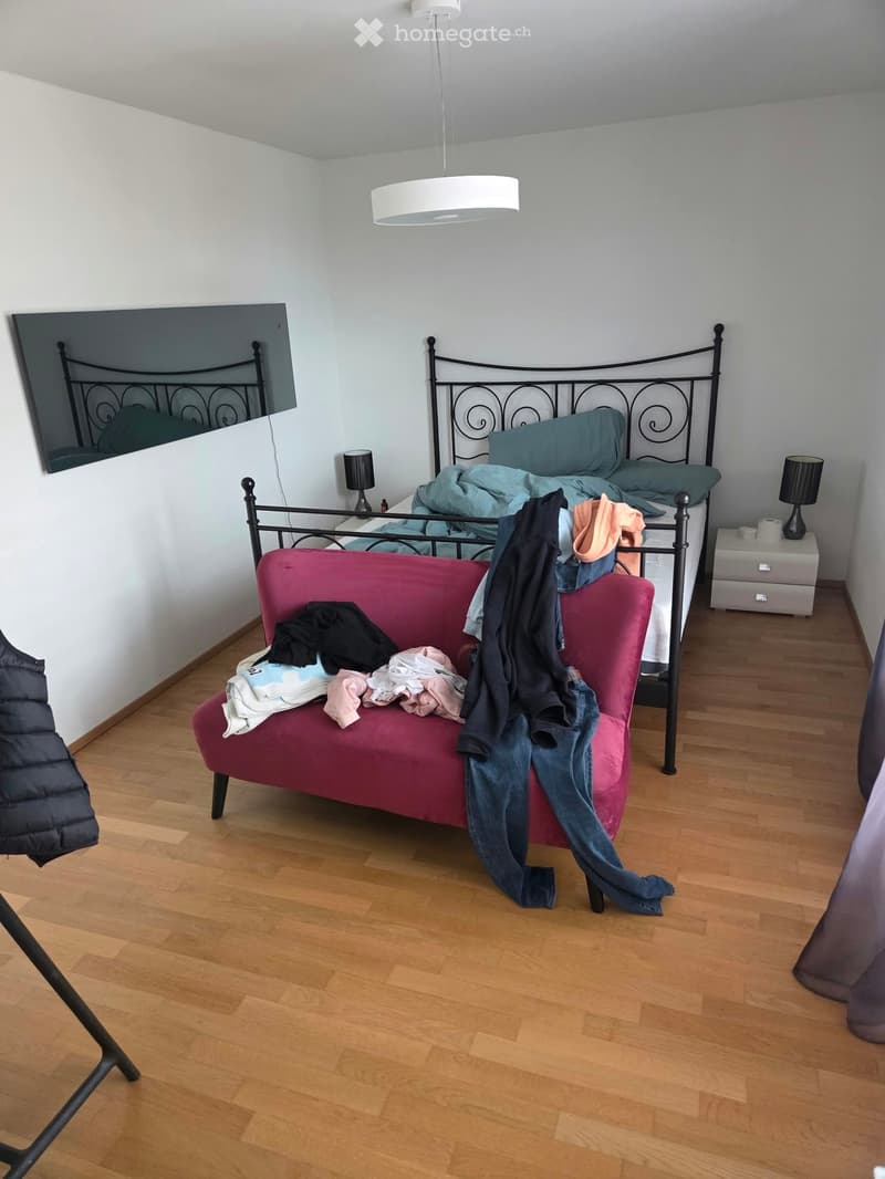 A bedroom with a wooden floor, a bed with a black metal frame and teal bedding, a red sofa with folded clothes, a mirror, and a pendant light.