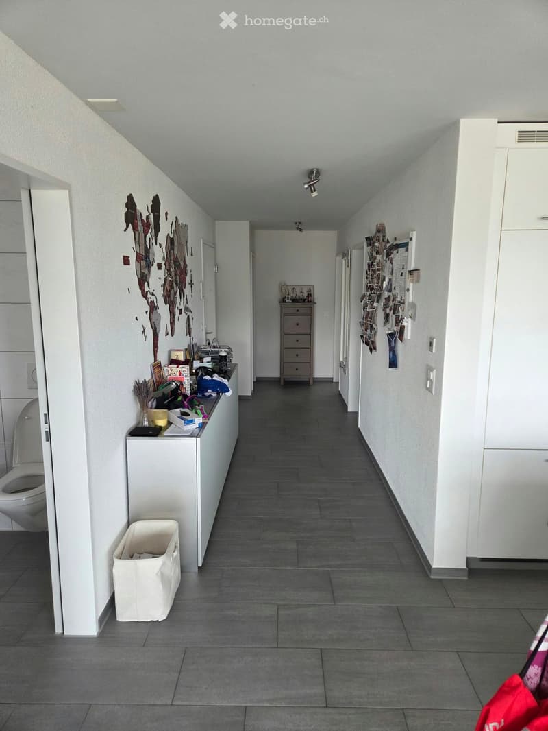A narrow hallway with grey tiled flooring, white walls, and several doors leading to different rooms. Visible items include a small cabinet, a trash bin, and a partially visible toilet in one of the rooms.