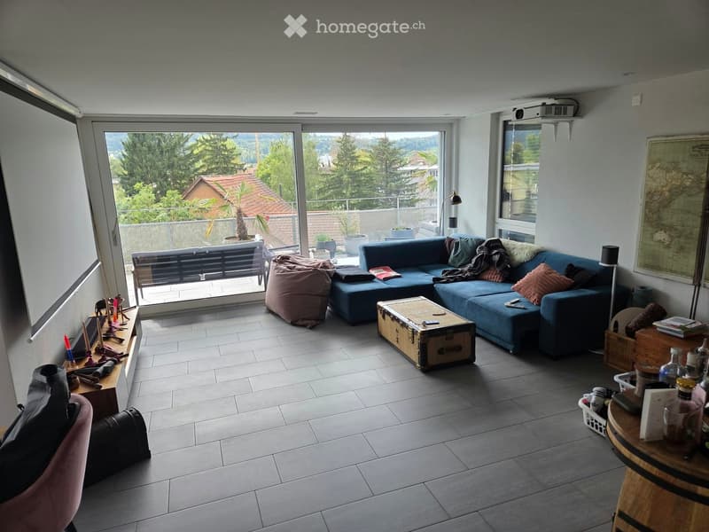 Modern living room with large windows offering a scenic view, featuring a blue sectional sofa, a wooden coffee table, a pink ottoman, and a round wooden table with various items.