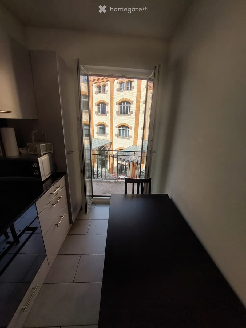 A small kitchen area with white cabinets, a microwave, and a dining table with a chair, opening onto a balcony with a view of a building.
