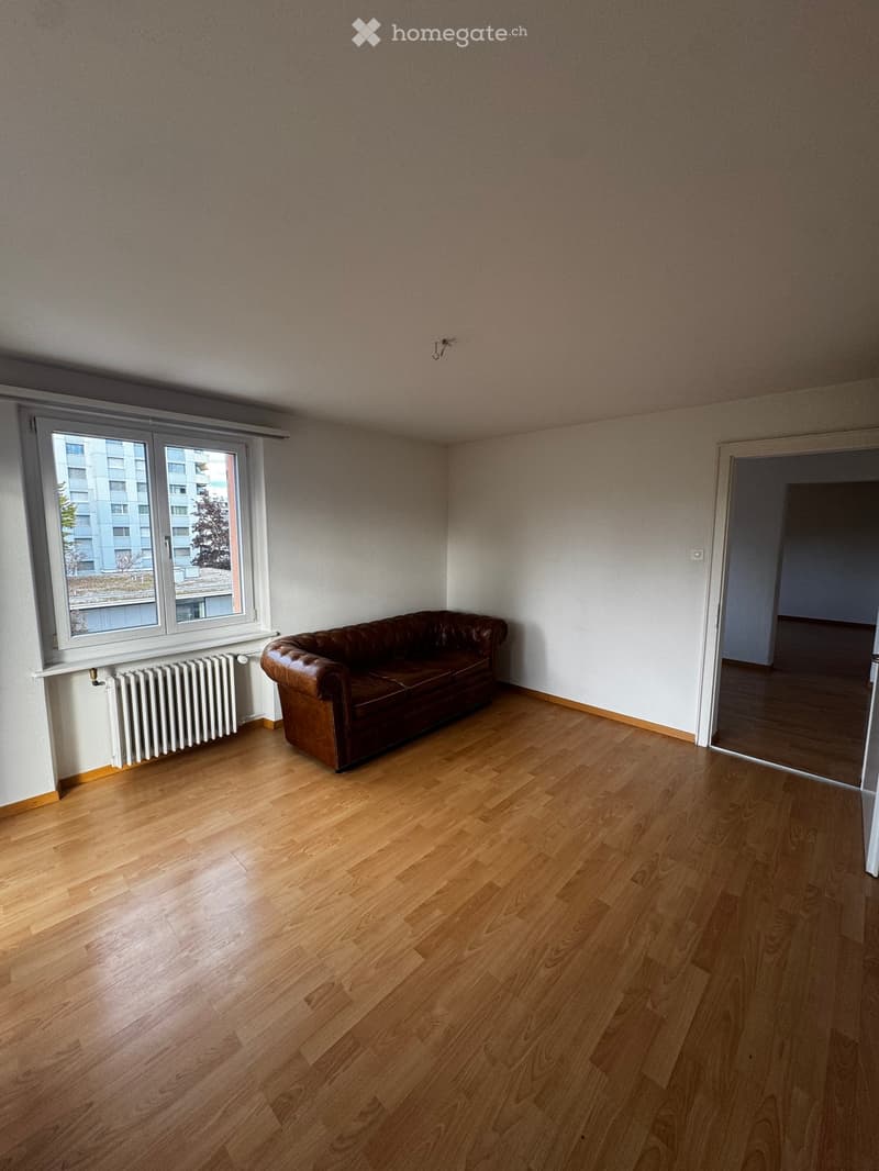 A furnished room with wooden flooring, a brown leather sofa, a window with a radiator, and a door leading to another room.