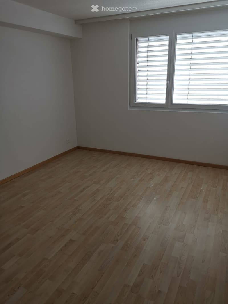 Empty room with wooden flooring and large window with blinds. Bare walls and no furniture.