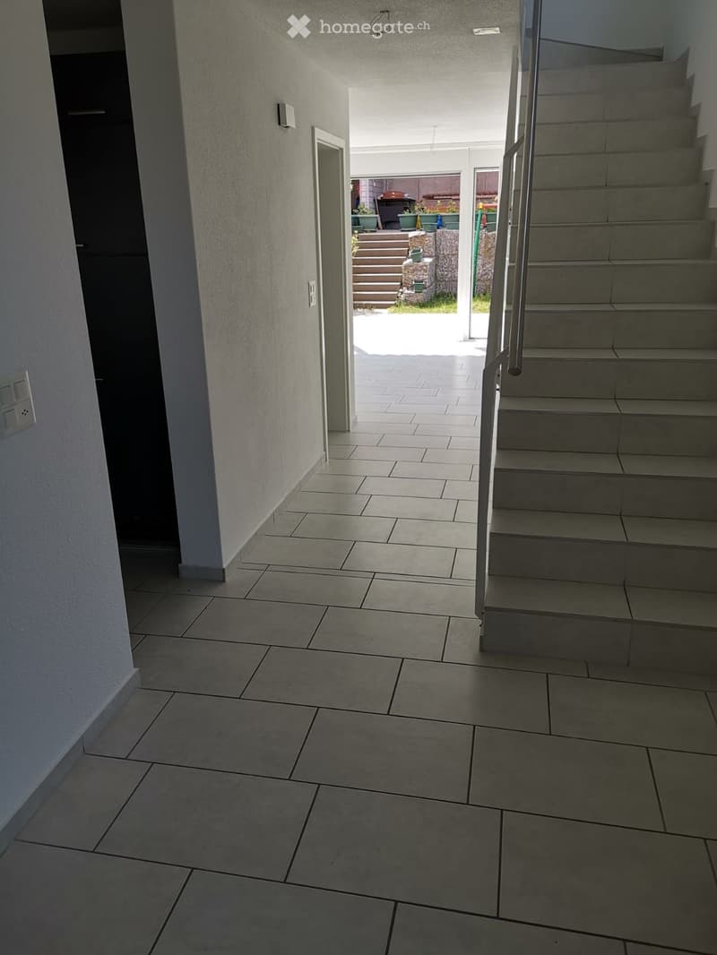 Hallway with tiled floor, staircase leading upwards, and an open doorway to an outdoor area with steps and potted plants.