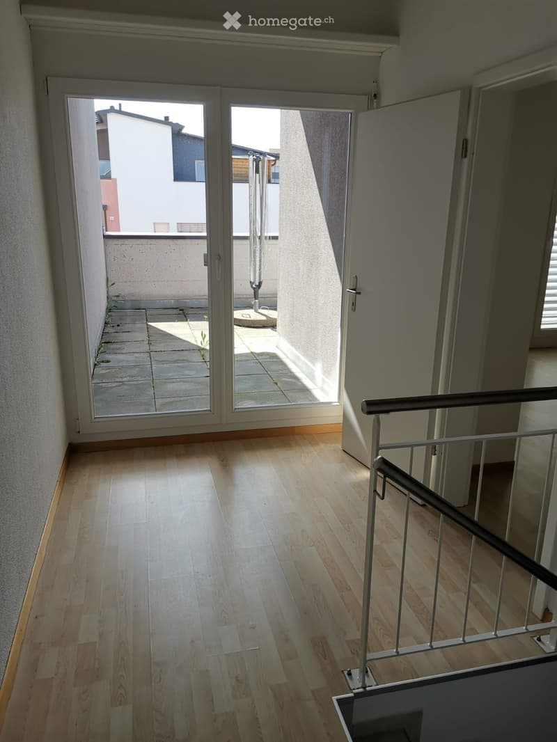 Empty hallway with wooden flooring, a staircase with a metal railing, and a large sliding glass door leading to a small outdoor area.