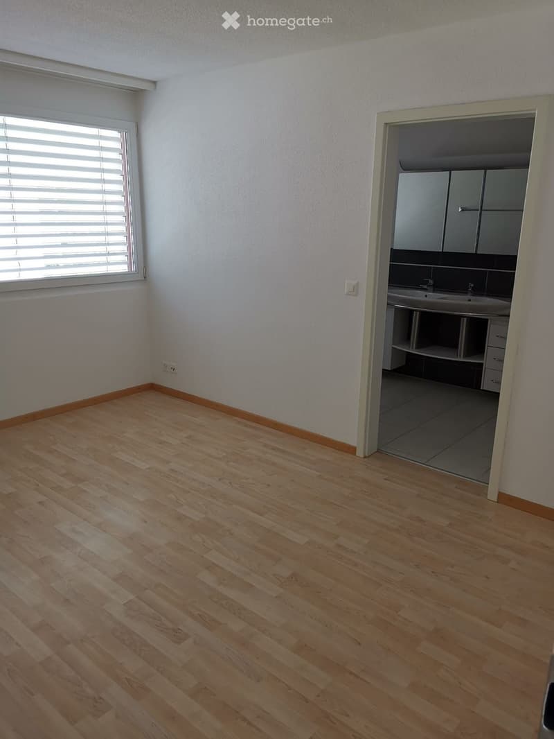 Empty room with light wooden flooring, white walls, and a window with blinds. An adjacent bathroom is visible with a sink, cabinets, and tiled flooring.