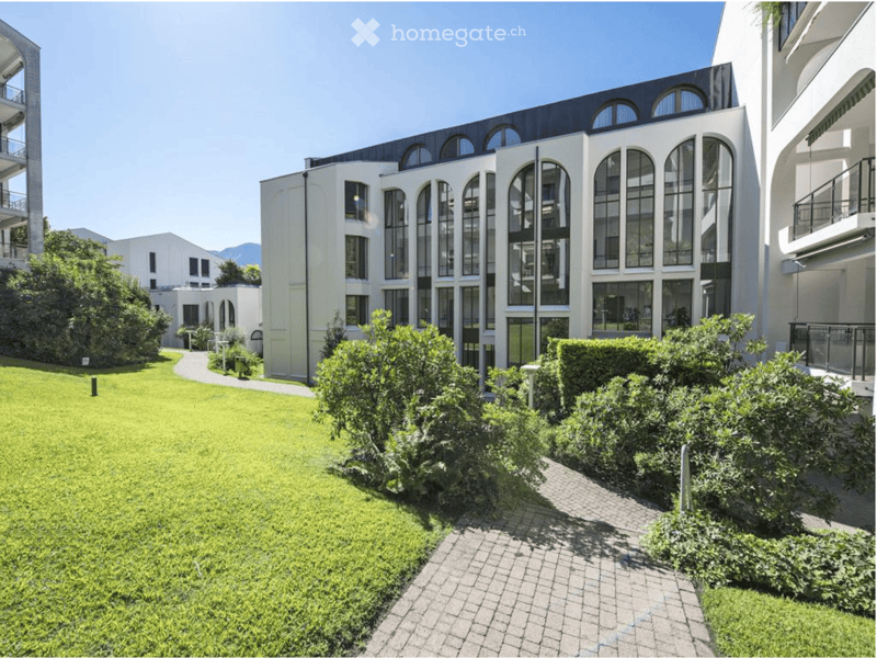 Well-maintained garden area with green lawn, trimmed bushes, and a paved pathway leading to a modern building with large arched windows and a sleek design.