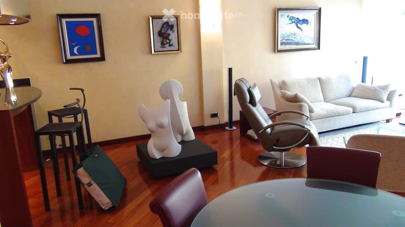 Living room with hardwood flooring, a grey sofa, a glass coffee table, a leather chair, a small black stool, and a white sculpture on a black base. Three framed artworks adorn the walls.