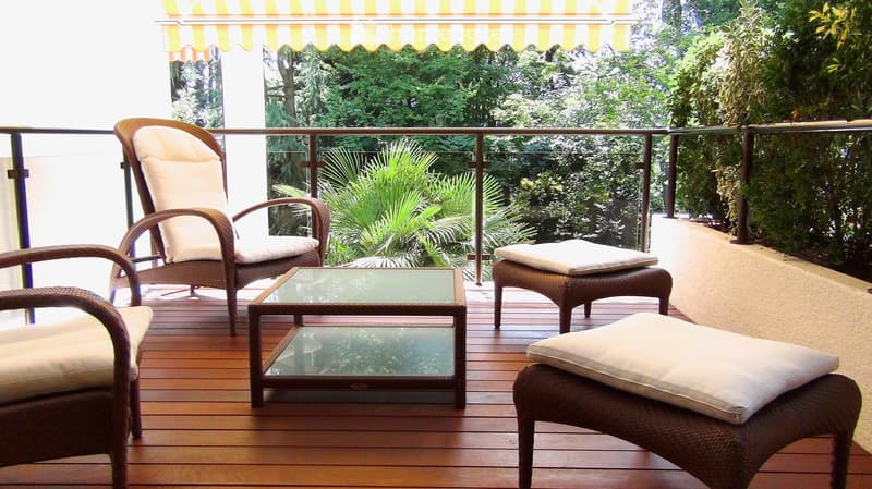 Balcony with wooden decking, wicker chairs, glass coffee table, and white cushions, overlooking a lush green garden.